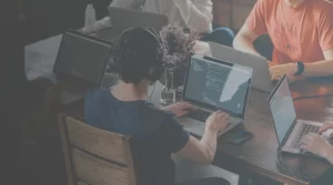 People collaborating at a wooden table with laptops and a headset-wearing coder in the foreground.
