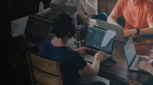 Person wearing headphones types on a laptop with code on the screen while teammates work nearby at a wooden table with laptops and a vase of flowers in the background.