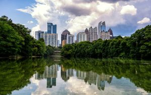 City skyline behind a tree-lined river, reflected in the calm water with clouds above.